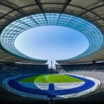 Panoramic view of an empty modern stadium with a circular glass roof opening and a green field.