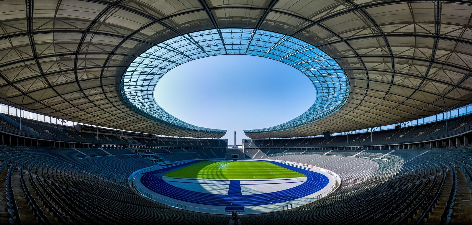 Альянц Арена - Черговий по Мюнхену Panoramic view of an empty modern stadium with a circular glass roof opening and a green field.