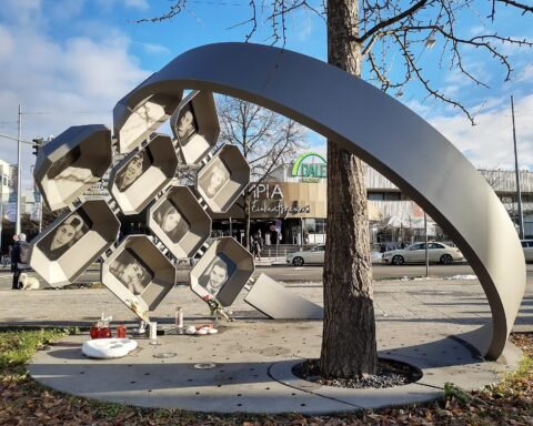 Outdoor sculpture: stacked hexagonal photo boxes surrounding a tree in a city plaza, containing black-and-white portraits and candles at the base indicating a memorial.