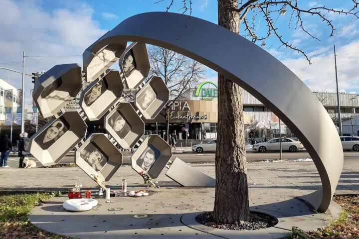 Outdoor sculpture: stacked hexagonal photo boxes surrounding a tree in a city plaza, containing black-and-white portraits and candles at the base indicating a memorial.