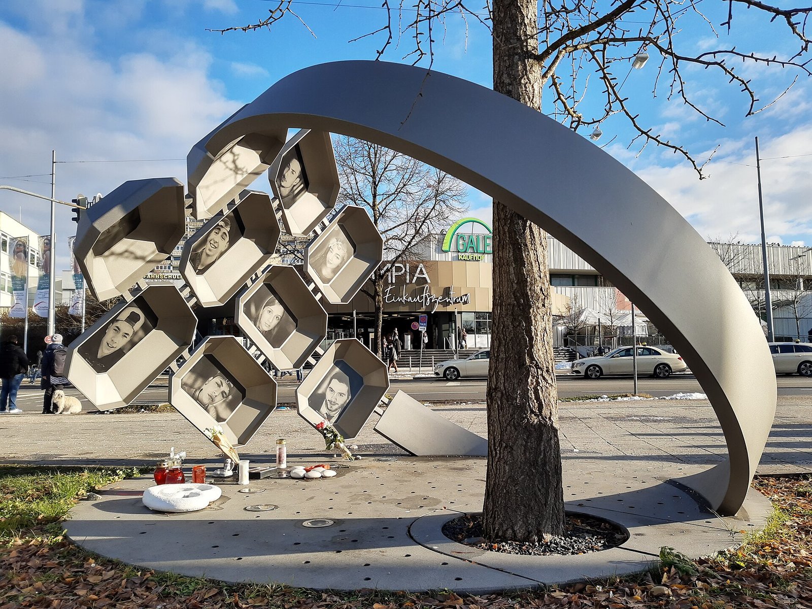 Теракт в ТЦ Олимпиа - On Duty of Munich Outdoor sculpture: stacked hexagonal photo boxes surrounding a tree in a city plaza, containing black-and-white portraits and candles at the base indicating a memorial.